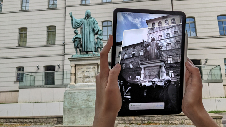 A person holds up a tablet in front of the Francke monument, on which a photo of the monument from 1990 can be seen. 