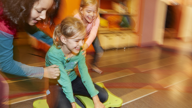 Three girls are playing. One sits on a board with rollers and is pushed by the other two. 