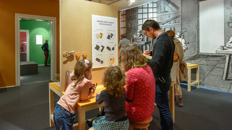 Eine Familie mit zwei Kindern baut in der Jahresausstellung eine kleine Camera Obscura mit einem Bastelsatz.