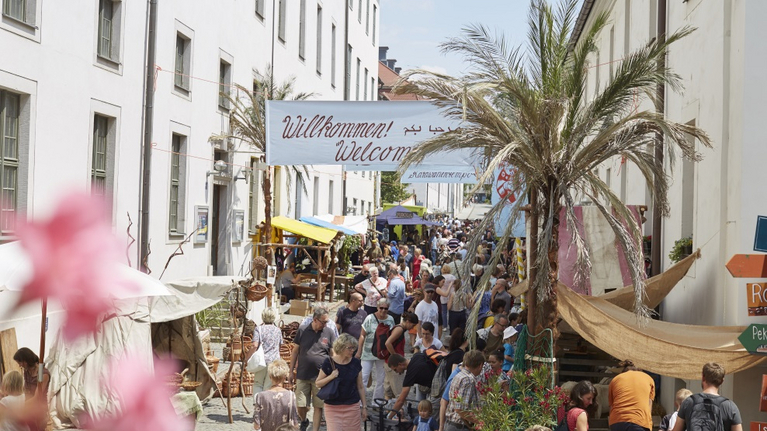 Sehr vielen Menschen sind zum Lindenblütenfest zu Besuch auf der Seidenstraße. Palmen säumen den Weg sowie viele Händler und Stände.