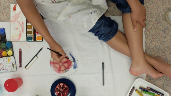 Ein Kind sitzt auf dem Boden und malt. Um es herum sind viele verschiedene Malutensilien ausgebreitet.
