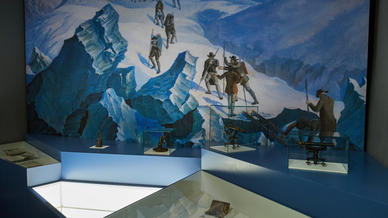 Blick in die Ausstellung auf ein wandgroßes Gemälde, auf dem Geologen einen verschneiten Gletscher besteigen.